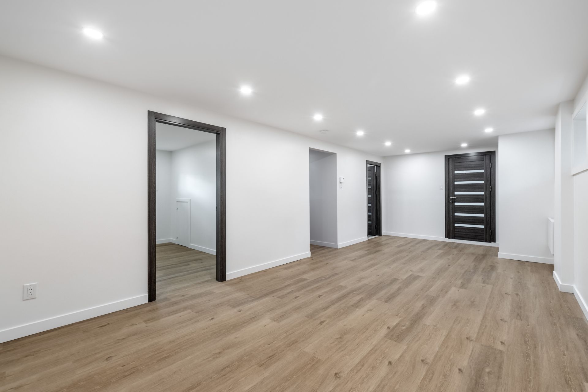 Empty, newly-renovated room with wood-look flooring, white walls, recessed lights, and black-framed doorways.