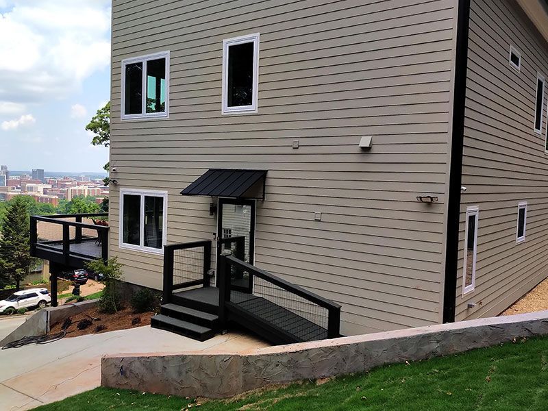 Two-story beige house with black trim, entry ramp, and awning, overlooks a city and a retaining wall.