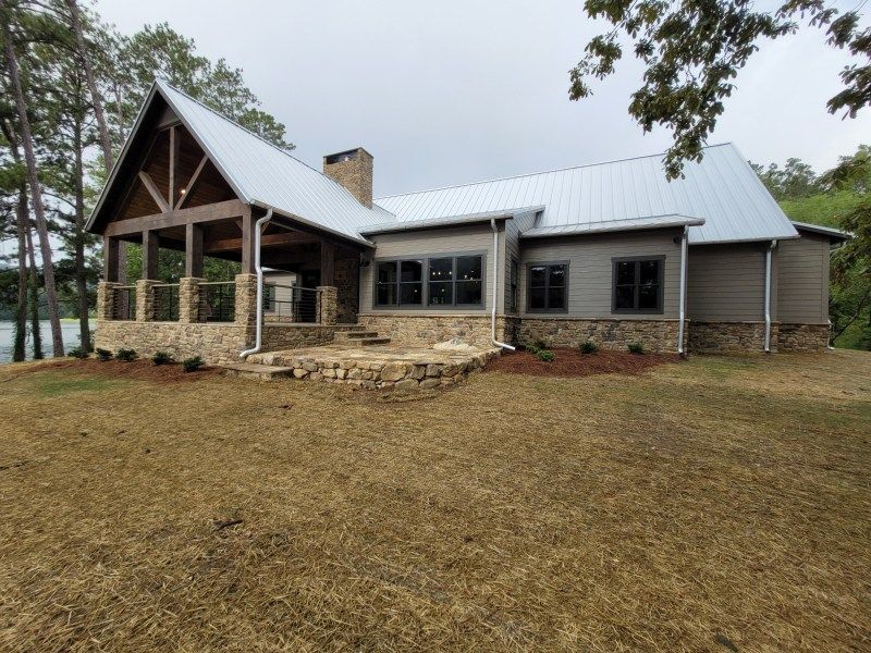 A gray house with a stone porch and roof, overlooking a lake, under a cloudy sky.