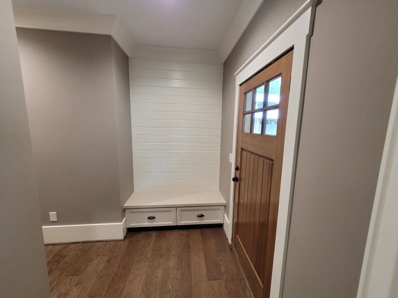 Mudroom with white shiplap wall, bench with drawers, brown door, beige walls, and wood flooring.