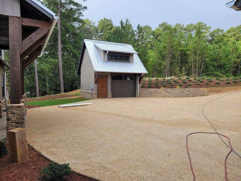 Gravel driveway leads to a light gray garage with a metal roof and a small window, set in a wooded area.