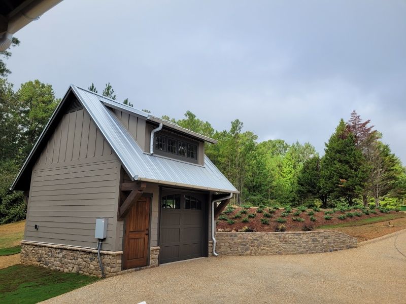 Small, modern garage with a metal roof, brown door, and stone base, in a grassy setting.