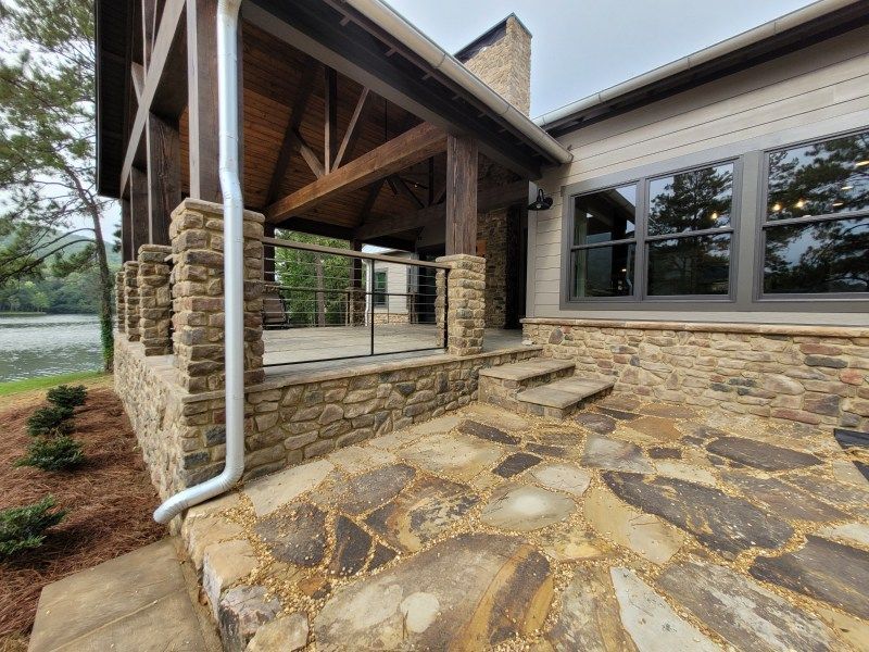 Stone patio with covered porch and stone columns next to a lake, with steps leading up to the door.