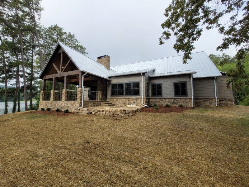 A house with a metal roof and stone accents sits on a grassy hill near trees and water.