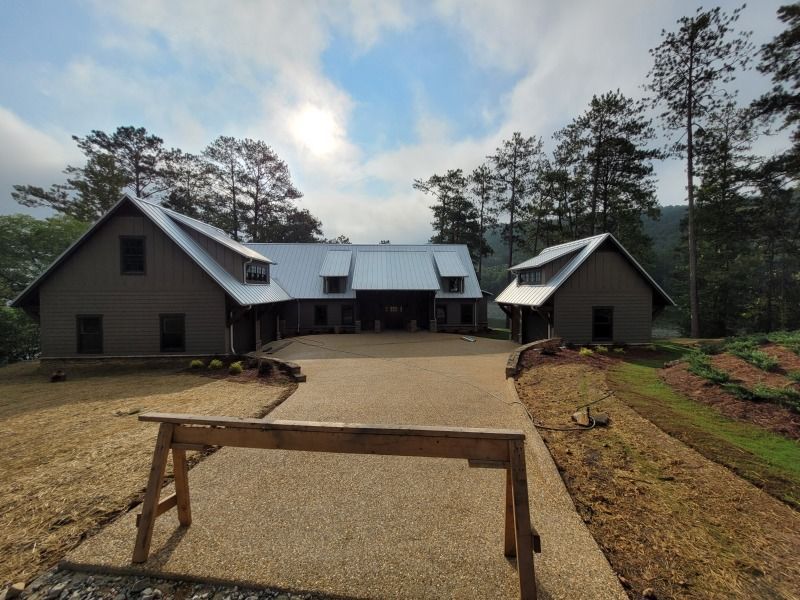 A gray house with a gravel driveway, surrounded by trees and a partly cloudy sky.