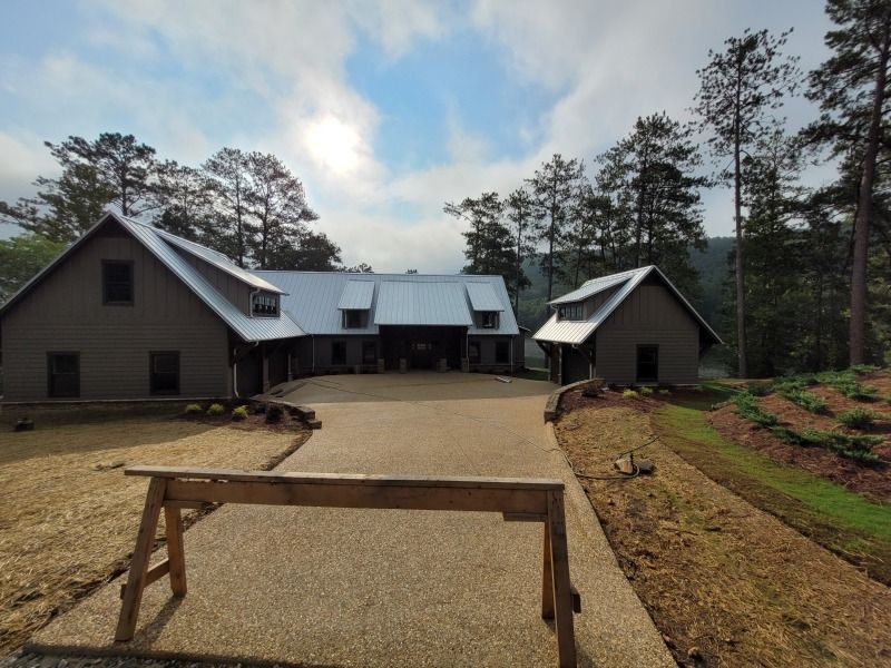 A large, gray house with a metal roof and a long driveway in a wooded area under a cloudy sky.