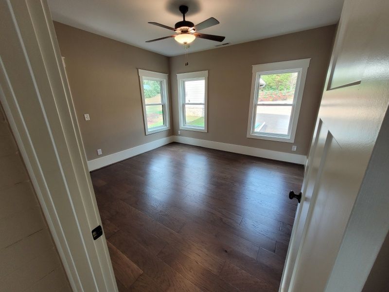 Empty room with wood floors, three windows, tan walls, and a ceiling fan.
