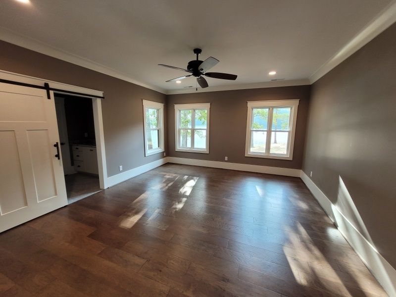 A spacious bedroom with wood floors, brown walls, white trim, windows, a barn door, and a ceiling fan.