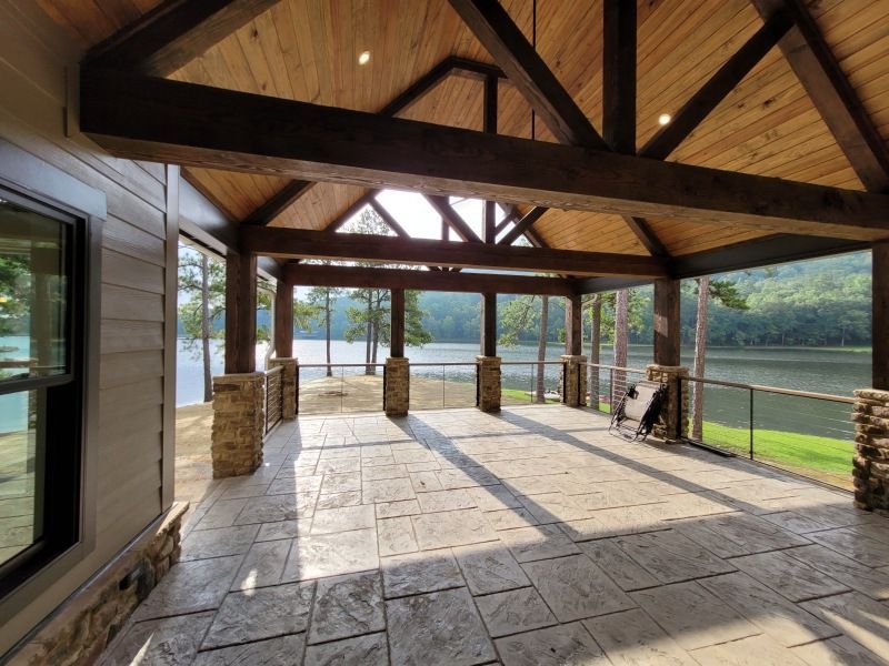 Covered outdoor patio overlooking a lake, featuring wood beams, stone columns, and a dog sitting near railing.