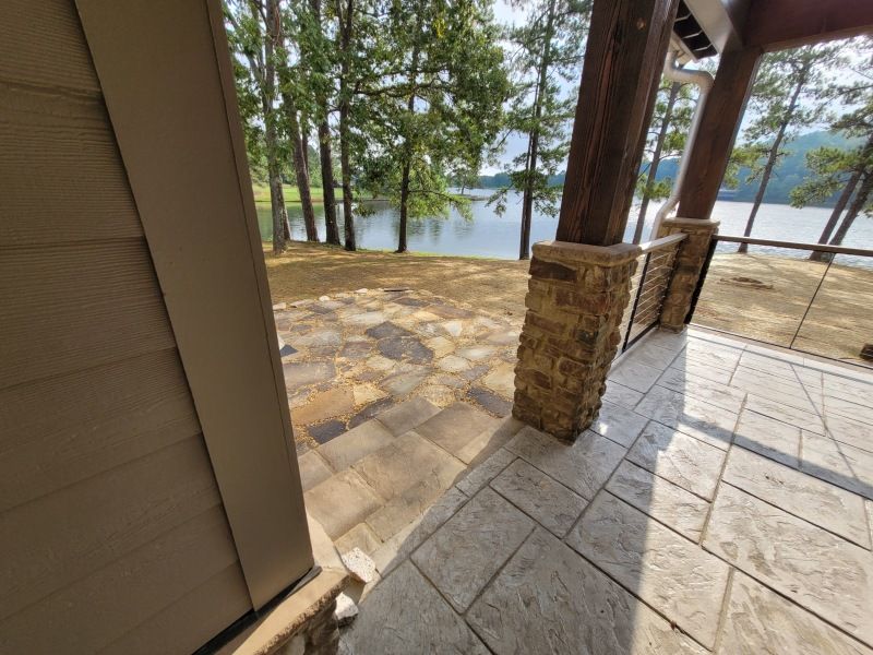 Patio overlooking lake, with stone columns, tan siding, and trees in background.