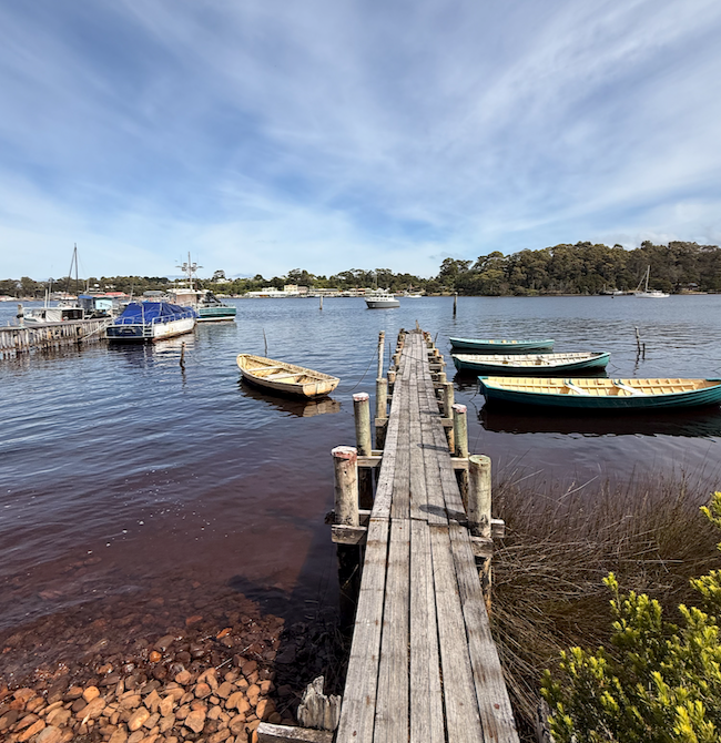 Wooden pier extends into a calm waterway with moored boats. Overcast blue sky and trees in the distance.