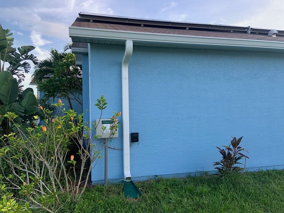 Blue house exterior with white gutter, greenery, and cloudy sky.