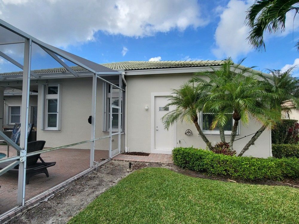 A light-colored house with a screened-in patio, surrounded by green grass and palm trees, under a blue sky.