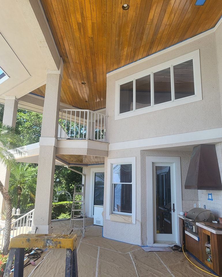 Exterior view of a house with wooden ceiling and white trim. Beige stucco walls, multiple windows, and a balcony.
