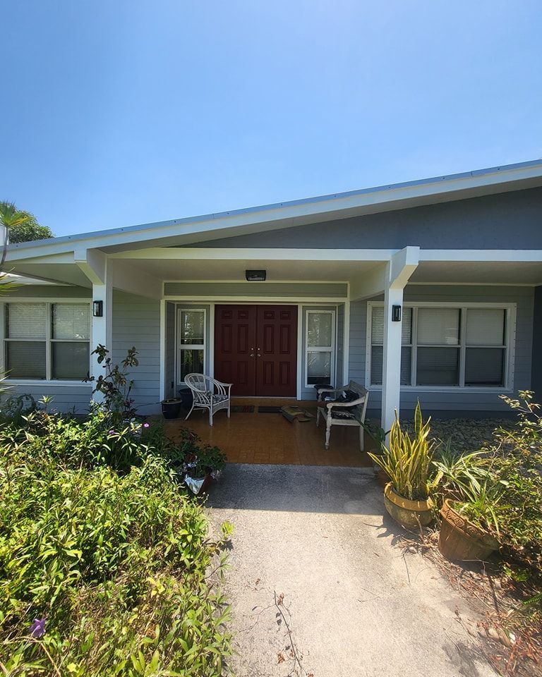 Grey house with covered entrance, red door, two white chairs, and pathway.