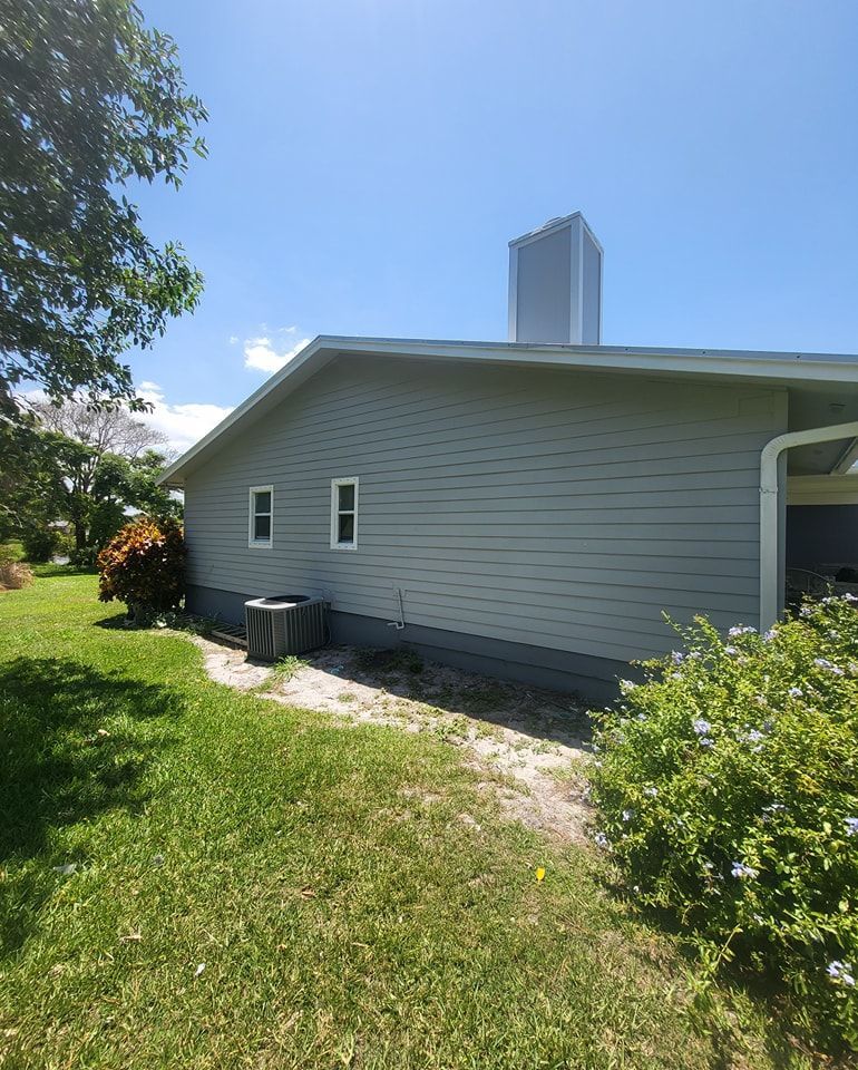 Side view of a house with light blue siding, two windows, and a chimney against a bright blue sky.