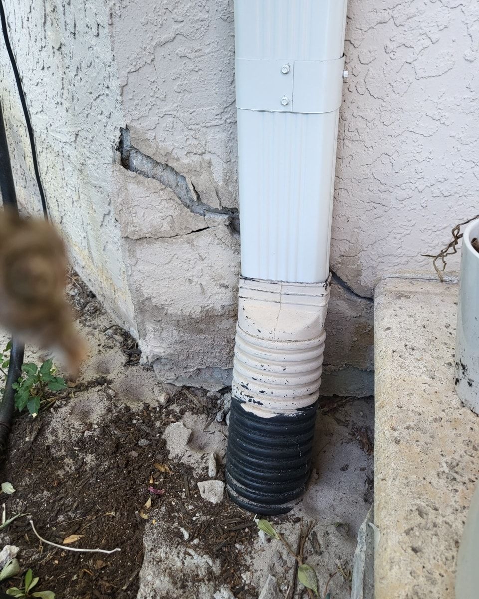 Downspout against a stucco wall; cracks are visible in the wall, debris at the base.