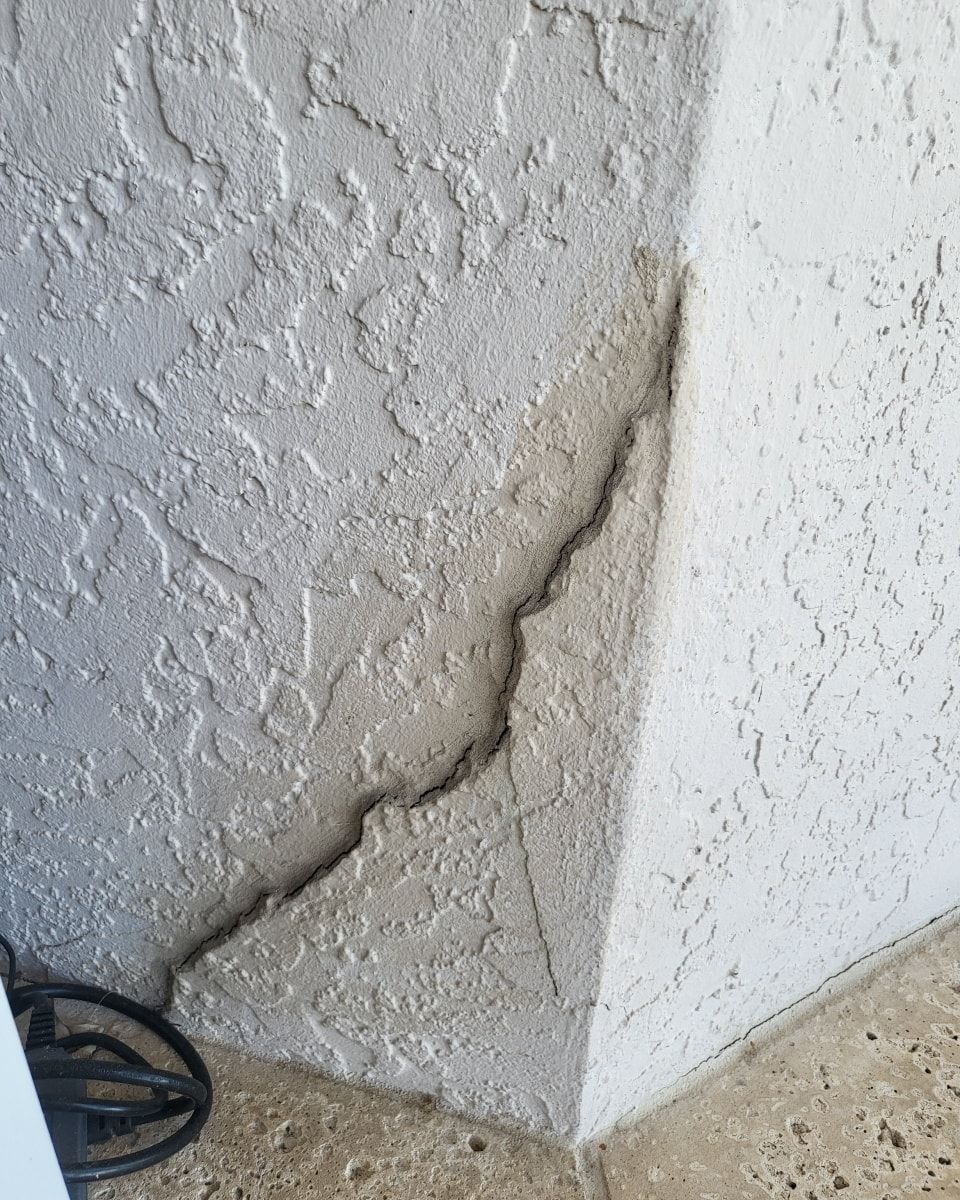 Cracked stucco corner of a building, with a jagged split running diagonally. Gray textured walls.