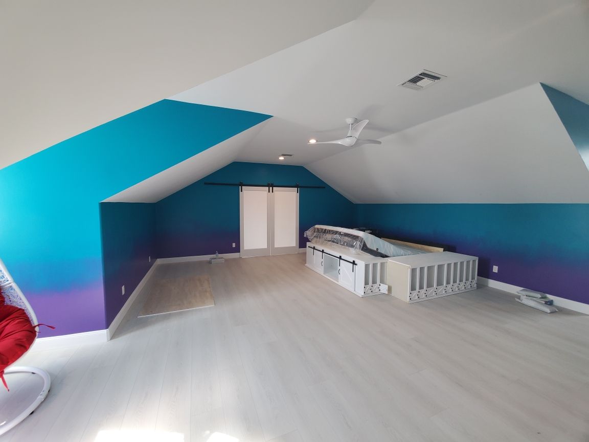 An attic room with gradient teal/purple walls, white ceiling, and pale wood floor. Large white doors in the back.