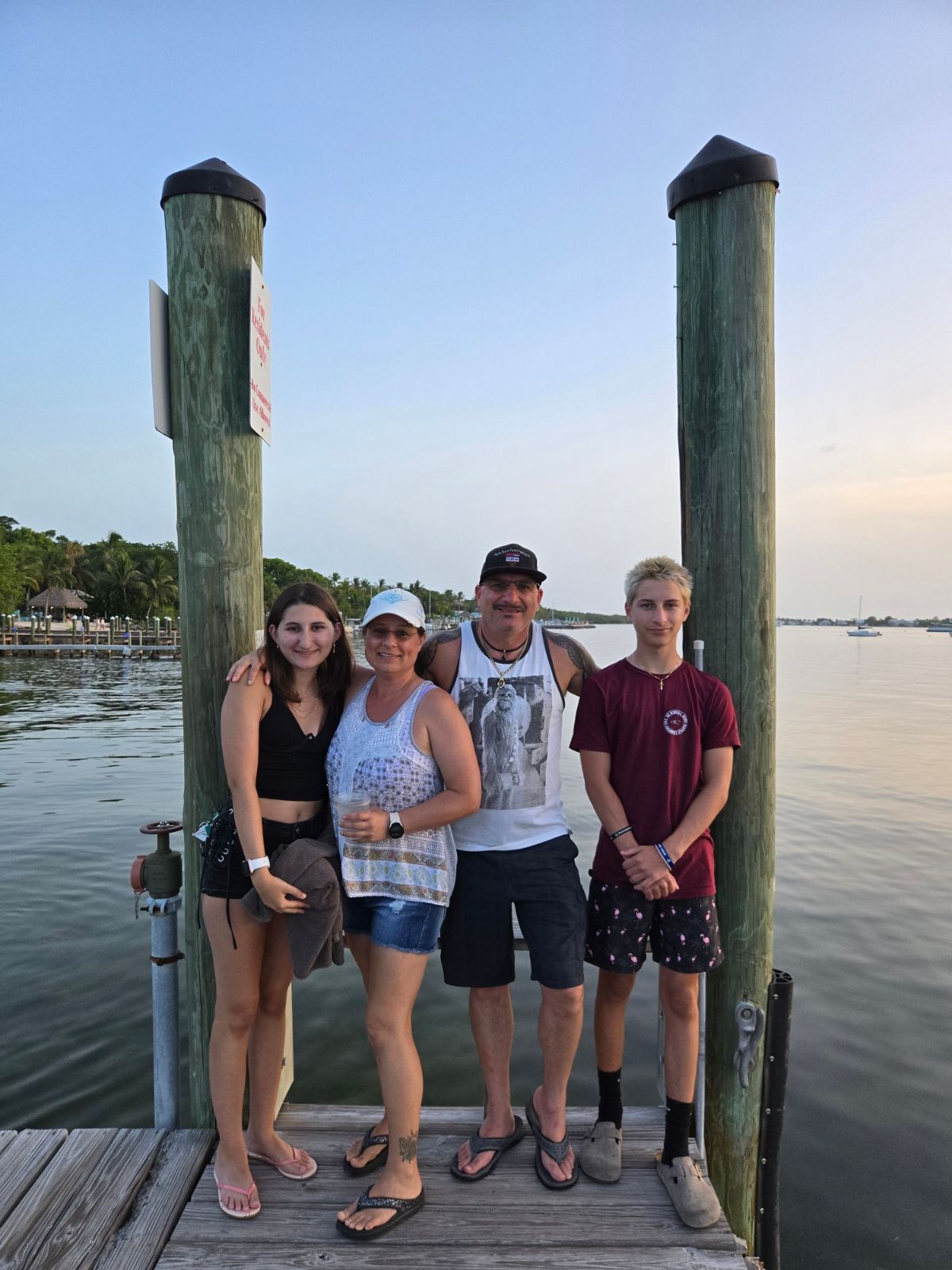 Family posing on a wooden dock at dusk, smiling. Two wooden poles frame the group near water.