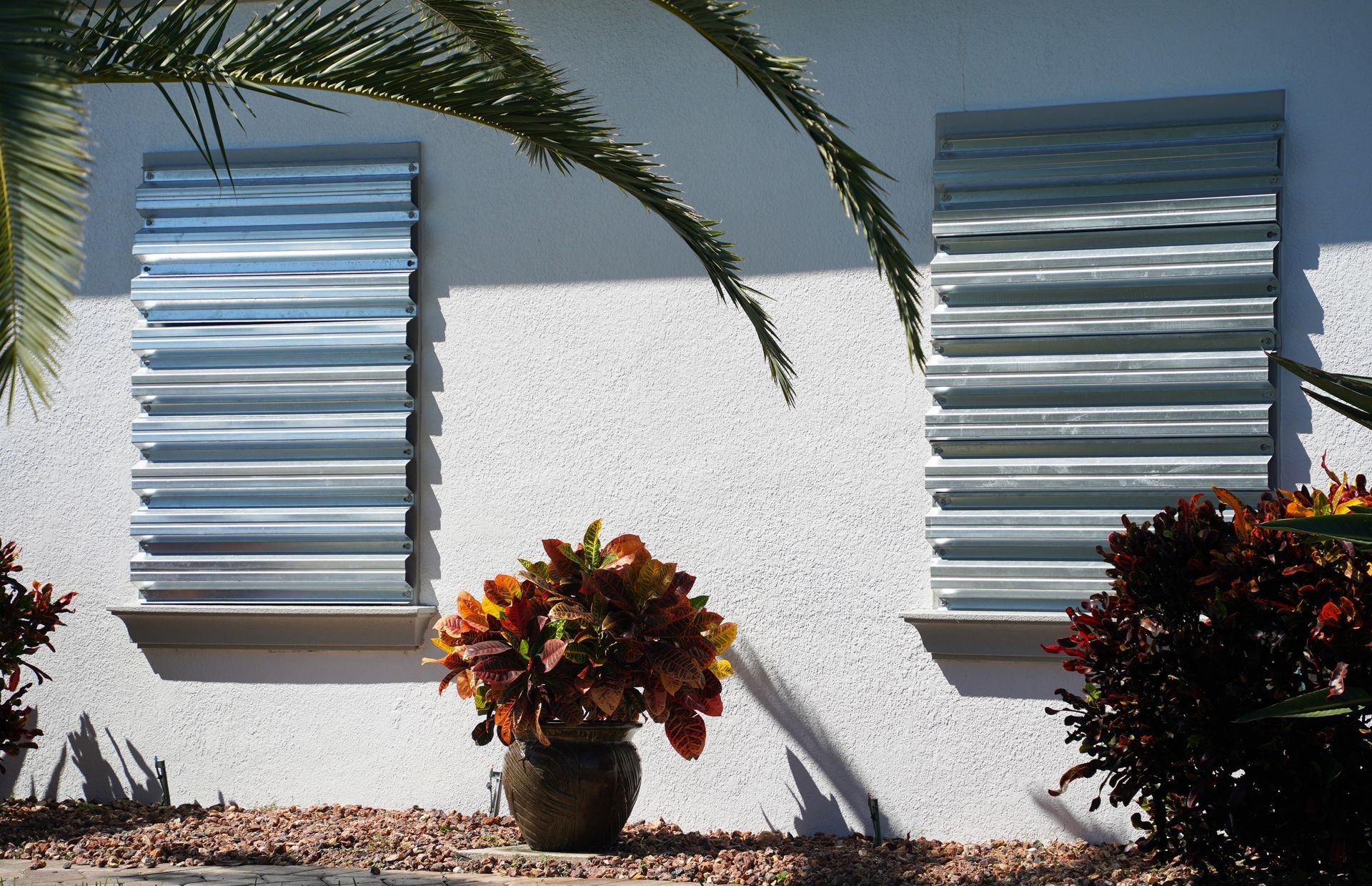 Windows with closed metal hurricane shutters on a white stucco wall, with a decorative flower pot in front.