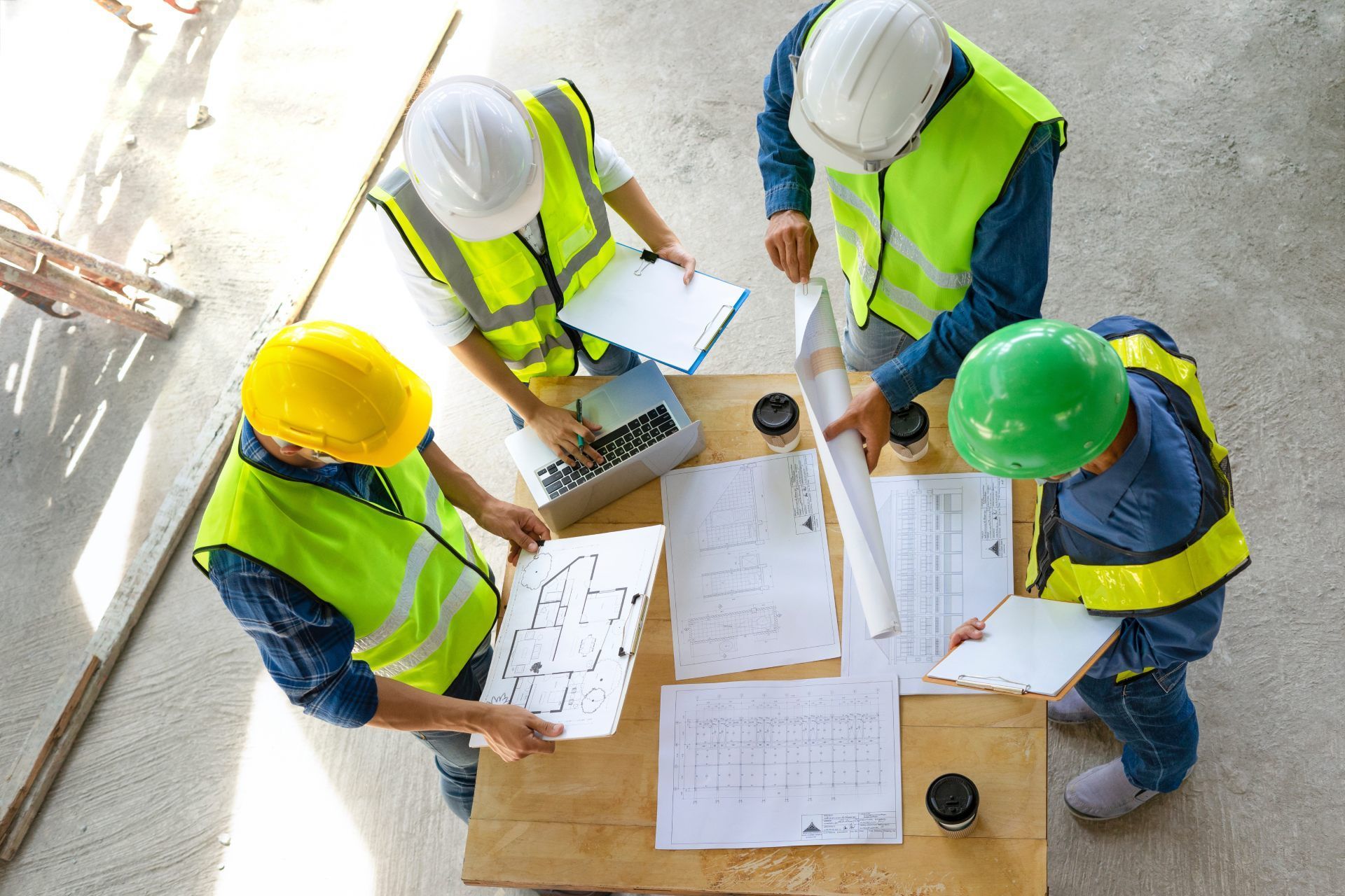Four construction workers in safety vests and hard hats reviewing blueprints around a table.