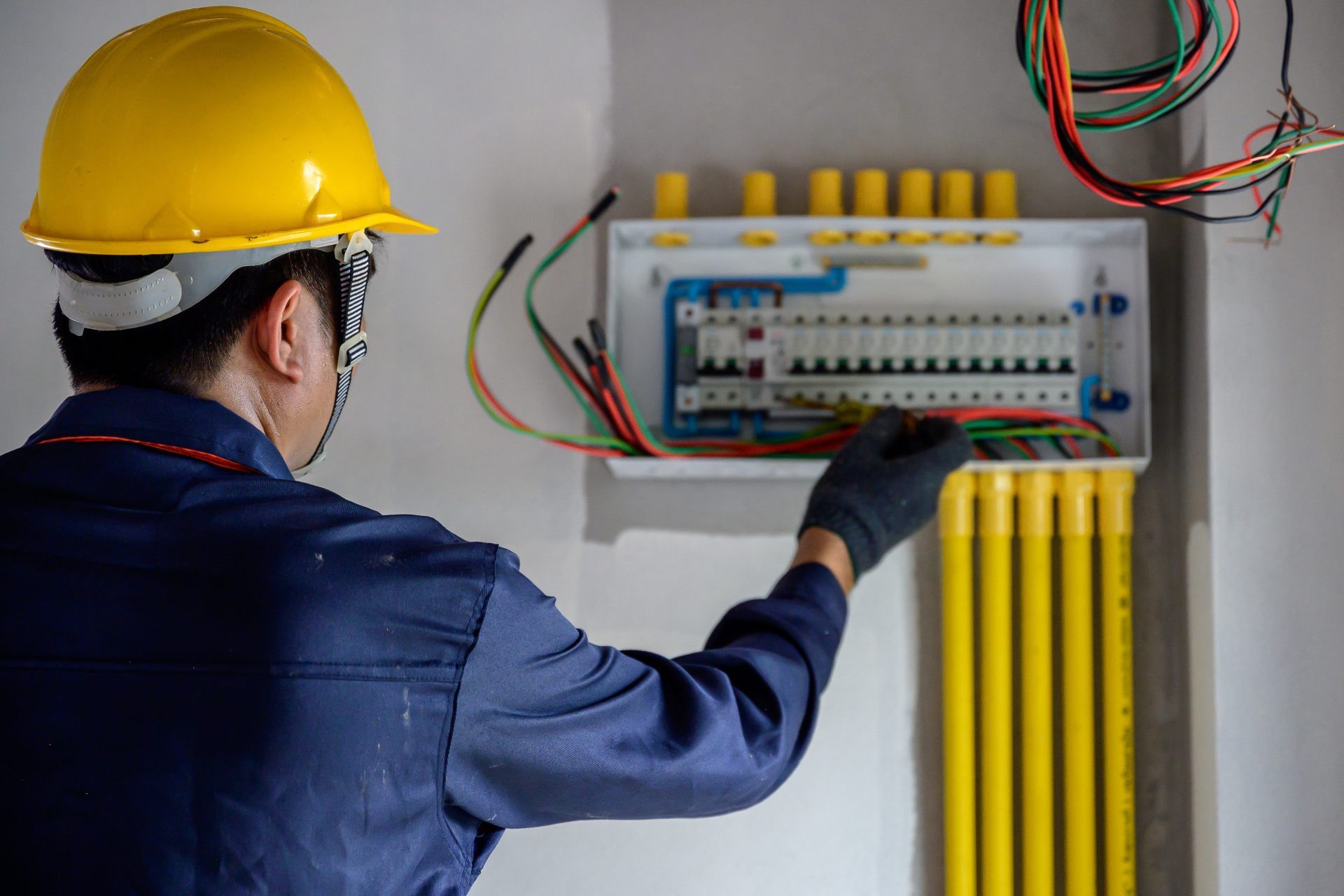 Electrician in blue coveralls and hard hat working on circuit breaker panel.