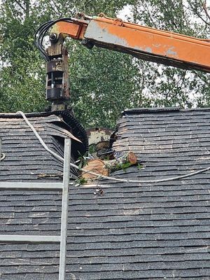 An excavator arm is demolishing a roof, creating a large hole. Orange arm, black roof, trees in the background.