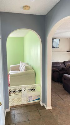 Arched doorway with green interior wall, white sofa, and a child barrier. Second arch leads to a living room.