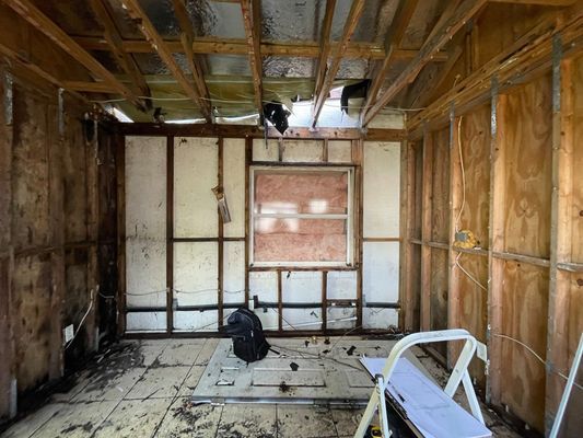 Interior view of a gutted room with exposed wooden studs, damaged walls, and debris on the floor; window in the center.