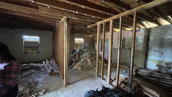 Interior of a room under renovation. Exposed framing and debris, with a person partially visible on the left.