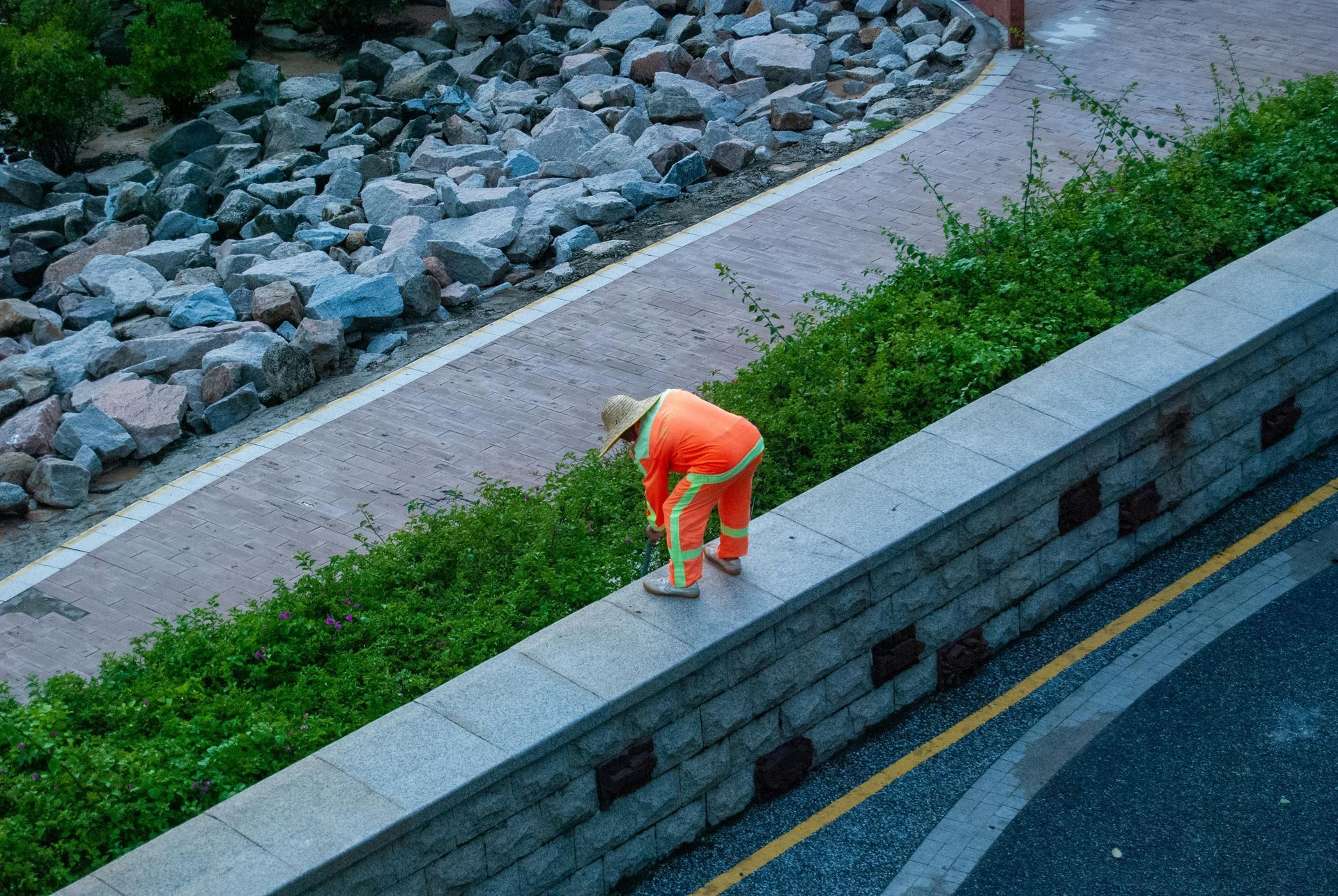 Person in orange work clothes standing on a stone wall, inspecting something. A path and greenery are nearby.