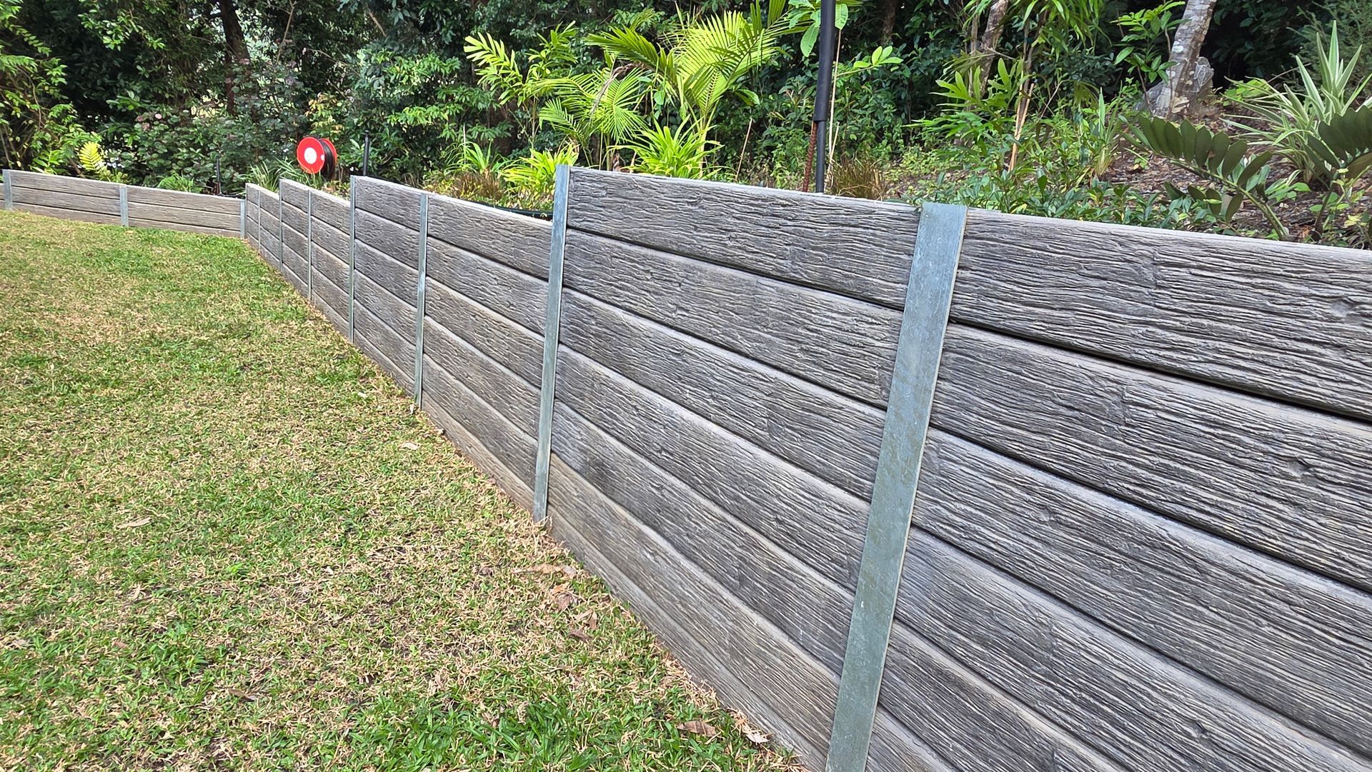 Sloped gray retaining wall with metal posts along a grassy hillside, trees in the background
