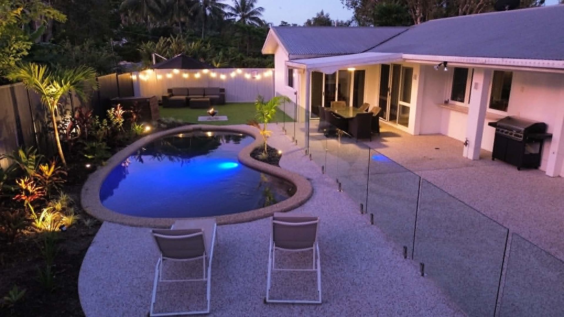 Backyard patio with glowing blue pool, lounge chairs, and a lit house at dusk