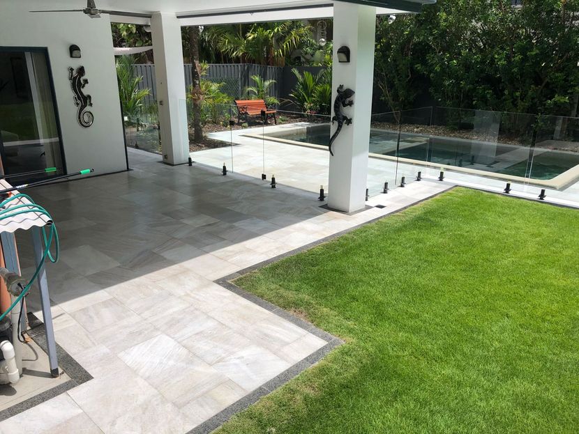 Patio with white pillars, gray tiles, green lawn, and a glass fence beside a poolside garden.