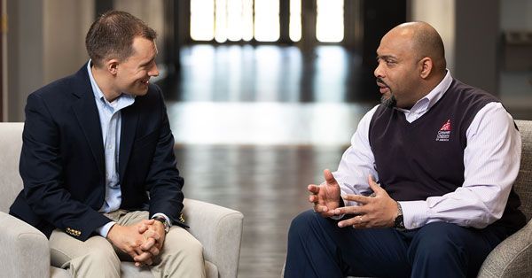 Two men are sitting on a couch talking to each other.