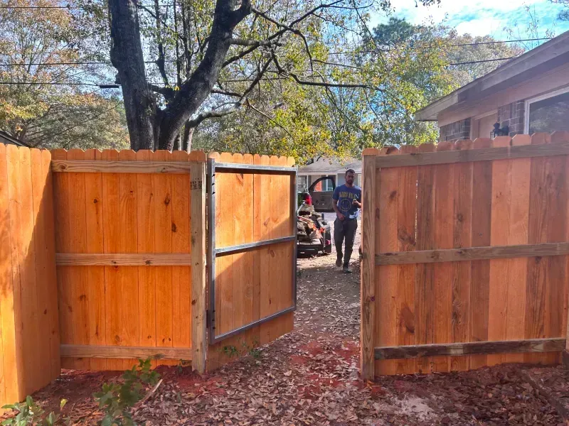 A man is standing behind a wooden fence with a gate open.