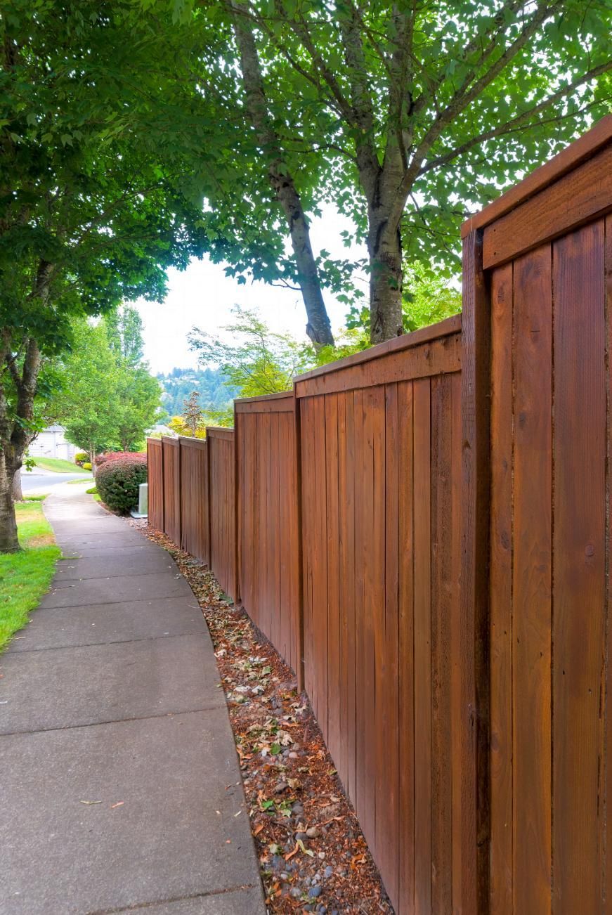 A wooden fence along a sidewalk with trees in the background.