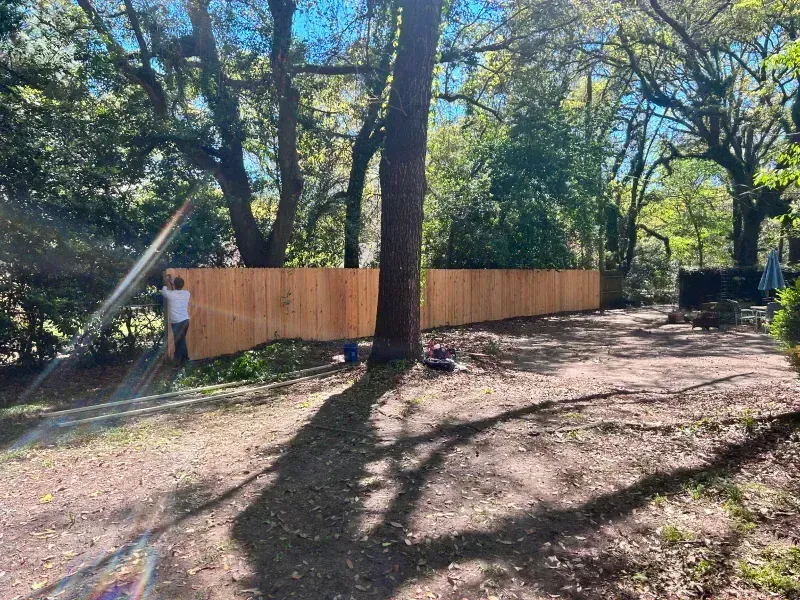 A man is standing next to a wooden fence in a yard surrounded by trees.