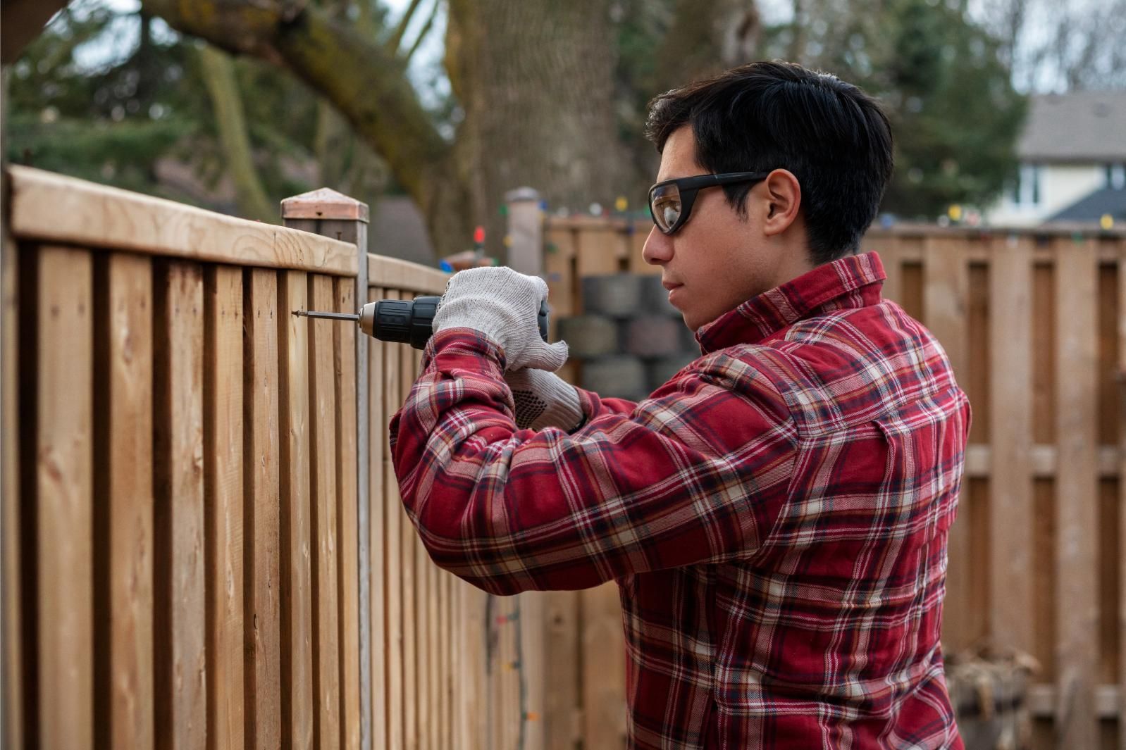 A man is using a drill to fix a wooden fence.