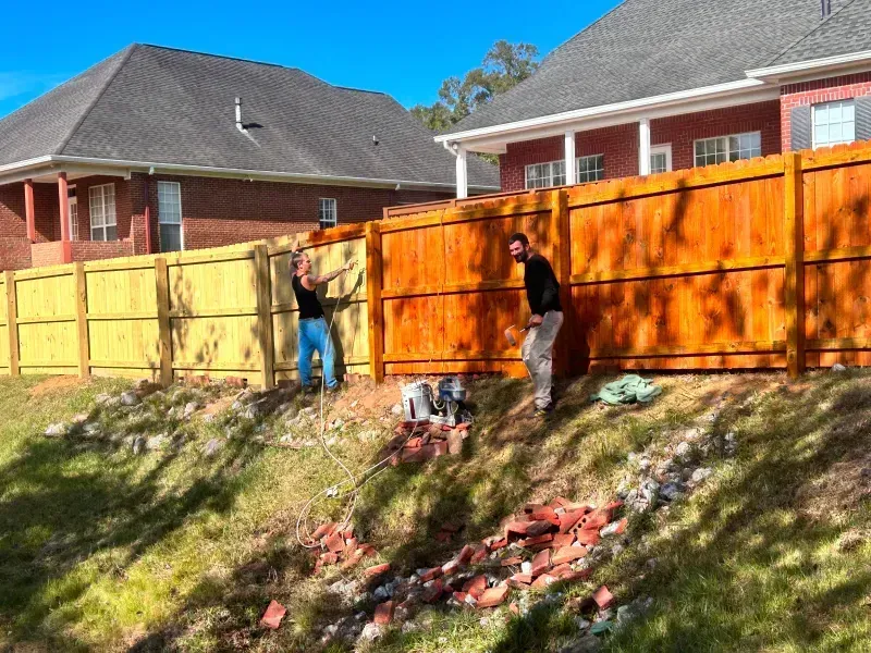 Two men are standing next to a wooden fence in front of a house.