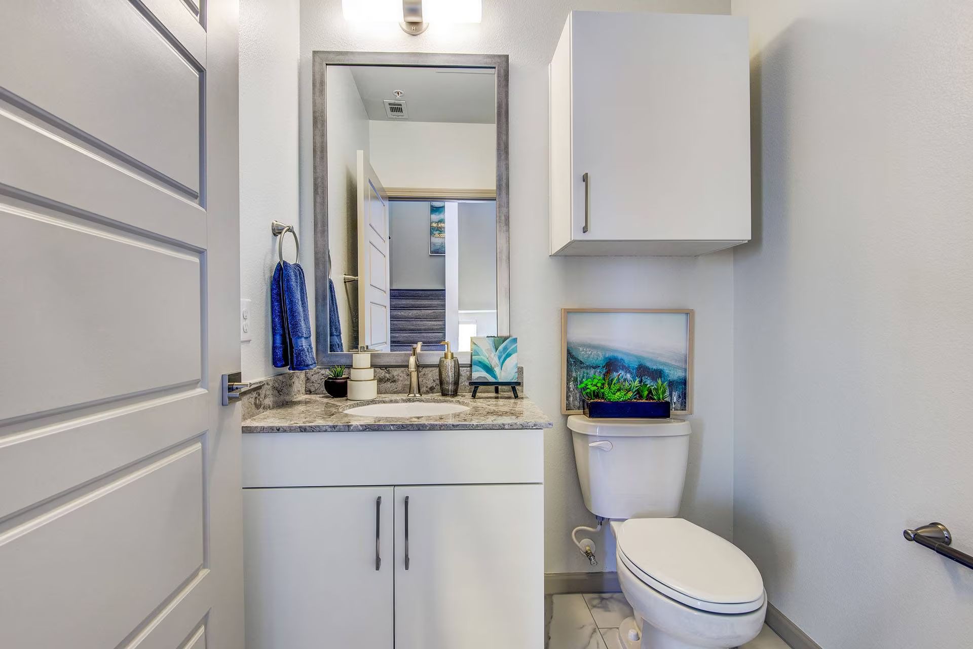 Small, modern bathroom with white cabinetry, toilet, and decorative painting. Silver framed mirror and marble countertop.