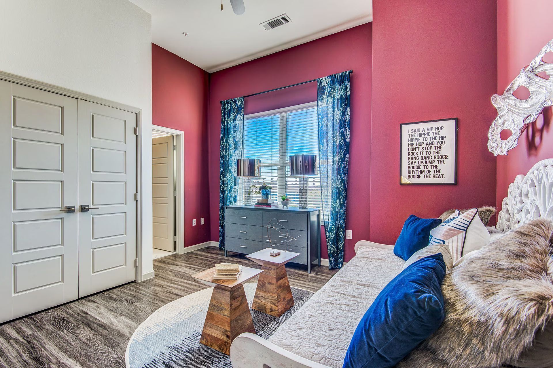 Bedroom with burgundy walls, white doors, gray dresser, blue curtains, and sofa with pillows.