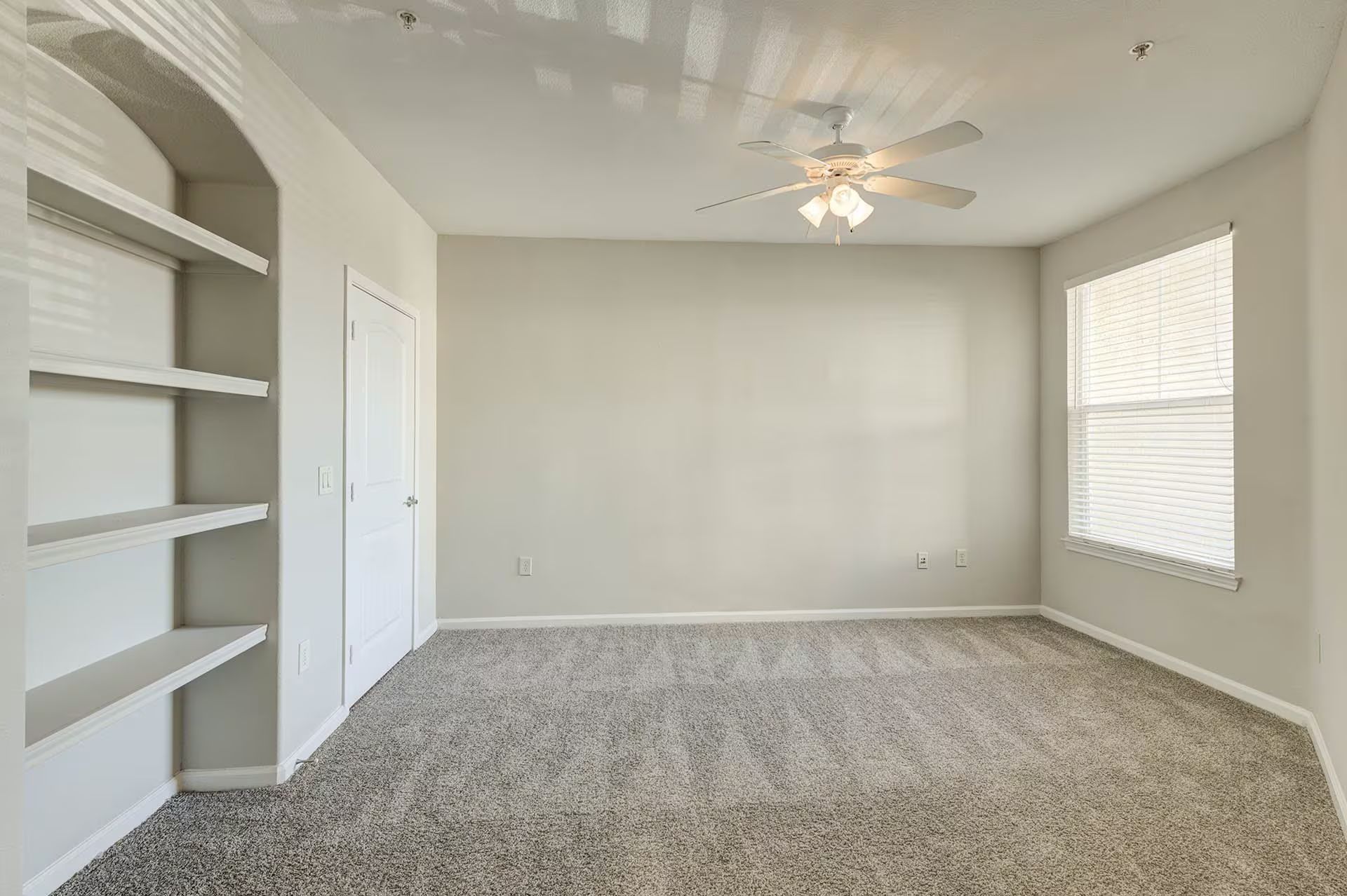 Empty room with grey carpet, built-in shelves, a window with blinds, and a ceiling fan.