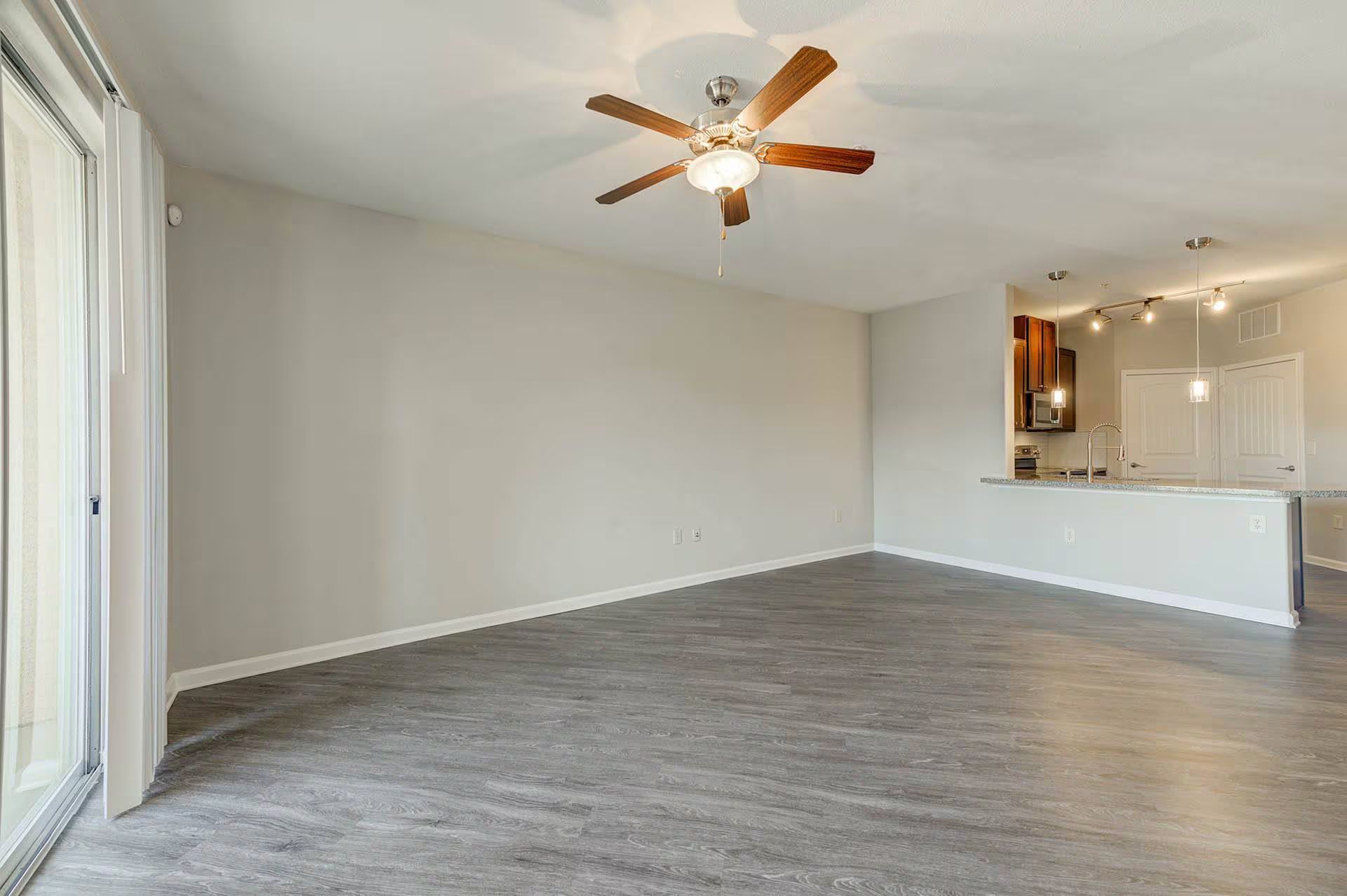 Empty living room with gray flooring, light gray walls, and a ceiling fan; kitchen in background.