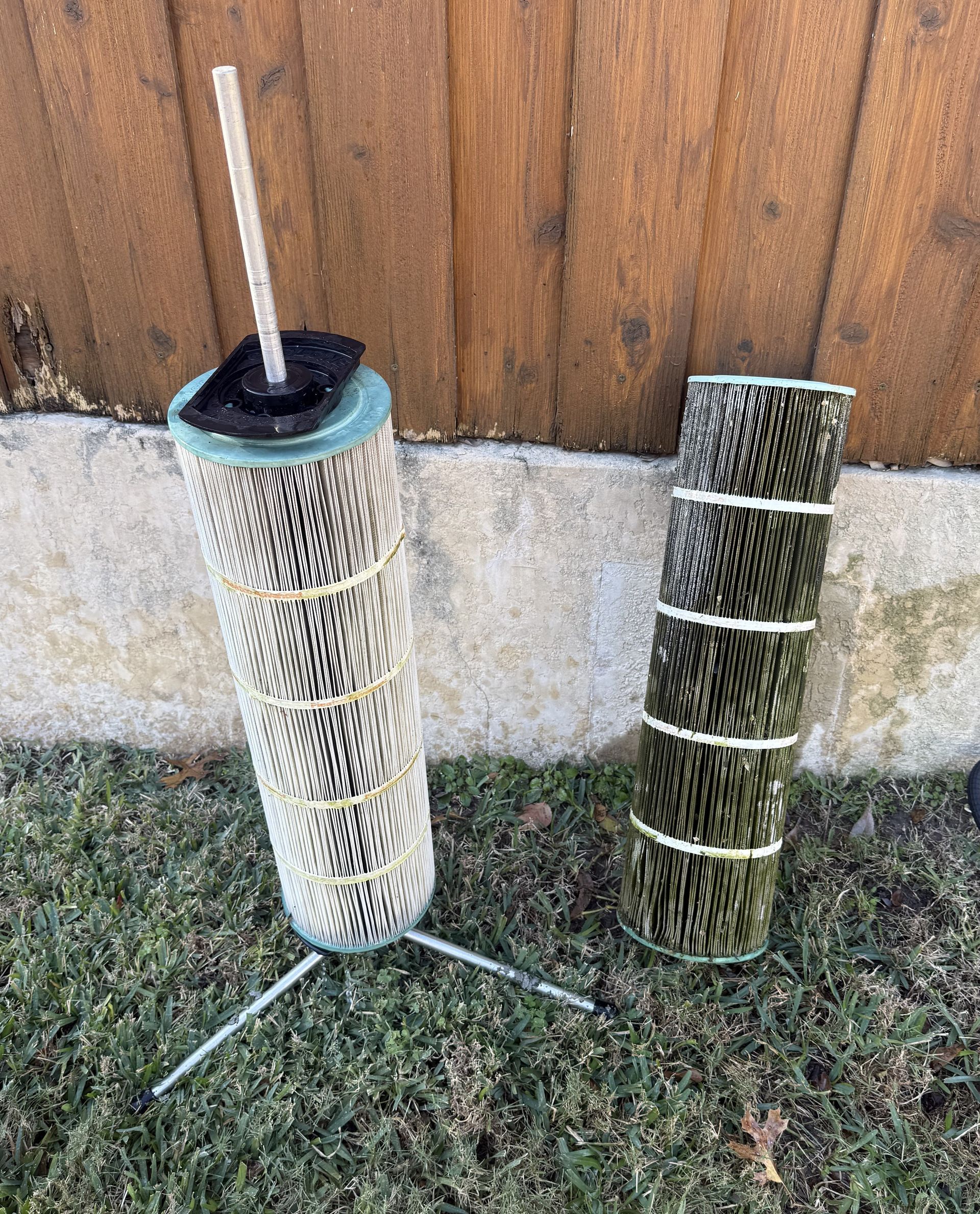 Two cylindrical air filters on grass, one with legs and a stick, the other lying down, against a wooden fence.