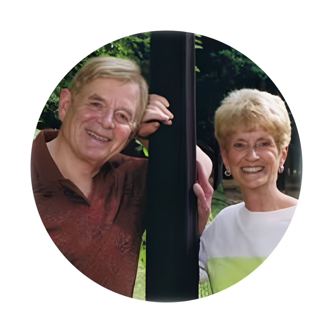 Elderly couple smiling, leaning against a dark post outdoors, surrounded by greenery.