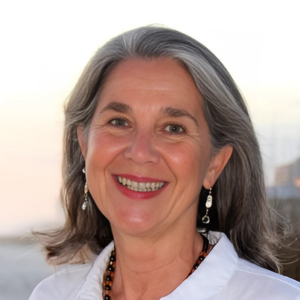 Woman with gray hair smiling, wearing white shirt, beaded necklace and earrings.
