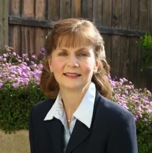 Woman in a navy blazer and white collar, smiles, in front of a wood fence and purple flowers.