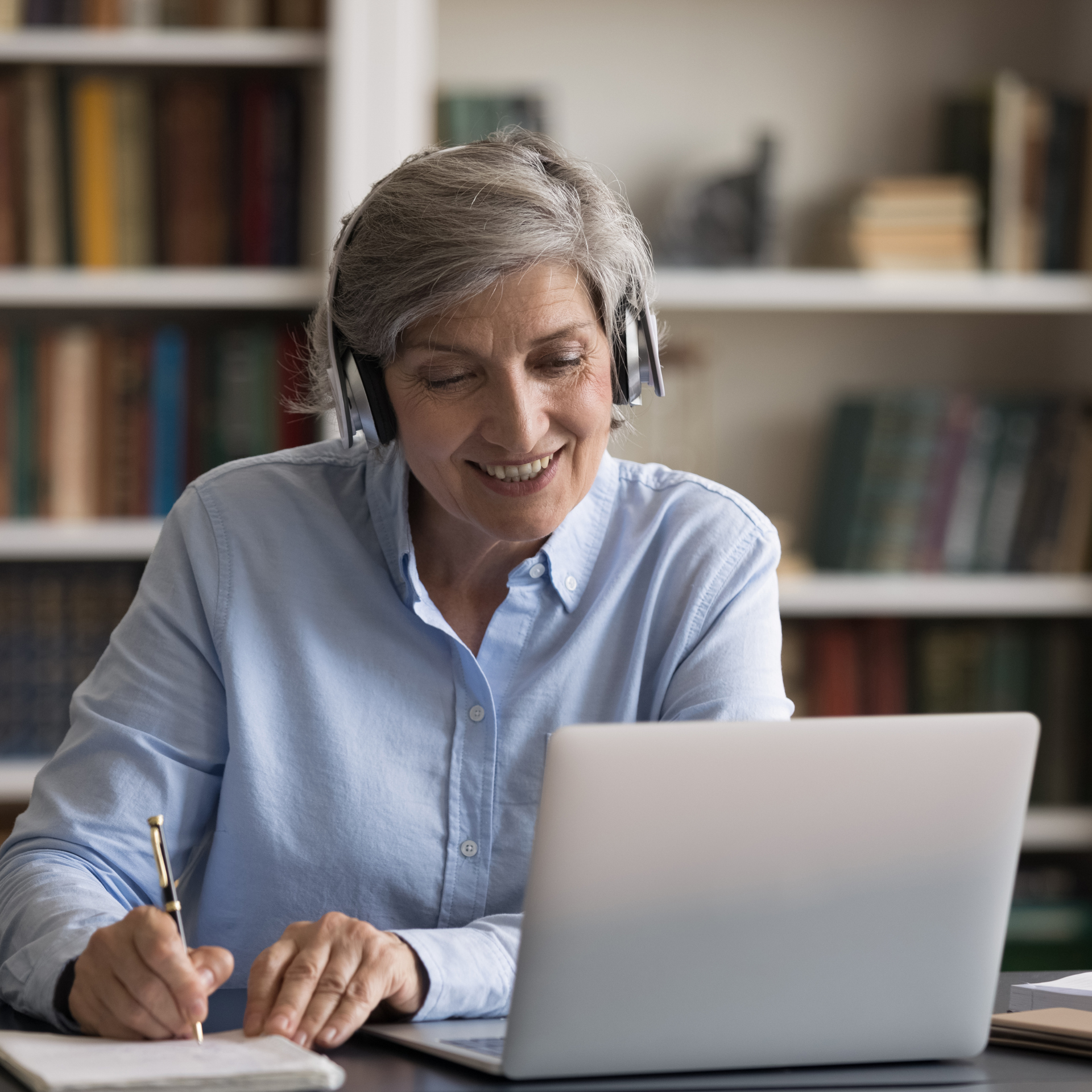 Older woman with headphones, smiling, taking notes while using a laptop in a library setting.
